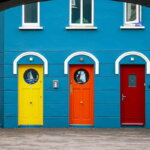 Drei farbige Türen in Gelb, Orange und Rot an einer blauen Hausfassade in Irland – Symbolbild für die drei verwandten Begriffe Ausgabe, Aufwand und Kosten.