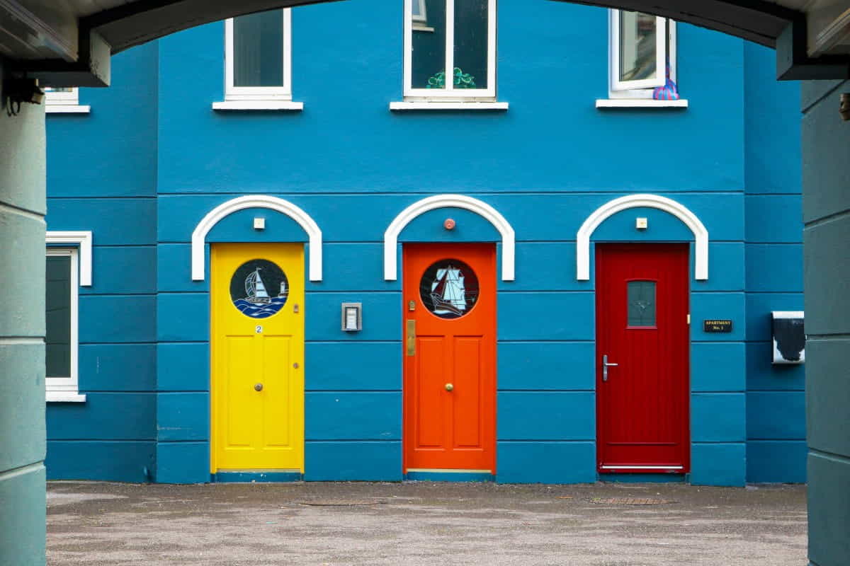Drei farbige Türen in Gelb, Orange und Rot an einer blauen Hausfassade in Irland – Symbolbild für die drei verwandten Begriffe Ausgabe, Aufwand und Kosten.
