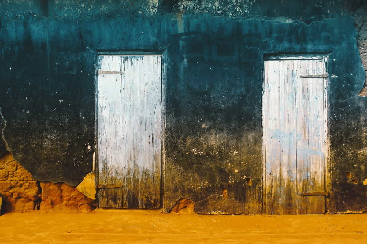 Zwei verwitterte weiße Holztüren in einer verfallenen blau-orangen Hausfassade – Symbolbild für den Unterschied zwischen Auszahlung und Ausgabe im Rechnungswesen.
