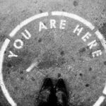 Standortanalyse / Symbolbild zur Standortanalyse: Blick auf Schuhe in einem markierten Kreis mit der Aufschrift „You are here“ – Standortwahl im Fokus.