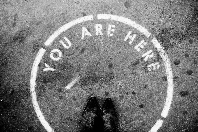 Standortanalyse / Symbolbild zur Standortanalyse: Blick auf Schuhe in einem markierten Kreis mit der Aufschrift „You are here“ – Standortwahl im Fokus.