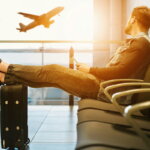 Checkliste Geschäftsreise / Mann sitzt auf Gang-Stuhl mit Füßen auf Gepäck und schaut auf das Flugzeug