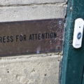 Messingschild mit der Aufschrift „Press for Attention" an einer Steinmauer neben einer verwitterten grünen Tür mit Klingel – Symbol für aktive Sichtbarkeit im Wettbewerb um Kundenaufmerksamkeit.