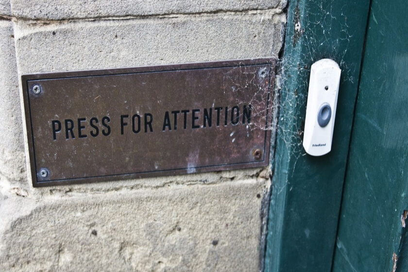 Messingschild mit der Aufschrift „Press for Attention" an einer Steinmauer neben einer verwitterten grünen Tür mit Klingel – Symbol für aktive Sichtbarkeit im Wettbewerb um Kundenaufmerksamkeit.