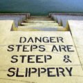 Scheinselbstständigkeit / Betontreppe führt ins Wasser, auf dem Boden steht in schwarzer Schrift: Danger - Steps are steep and slippery
