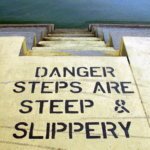 Scheinselbstständigkeit / Betontreppe führt ins Wasser, auf dem Boden steht in schwarzer Schrift: Danger – Steps are steep and slippery.