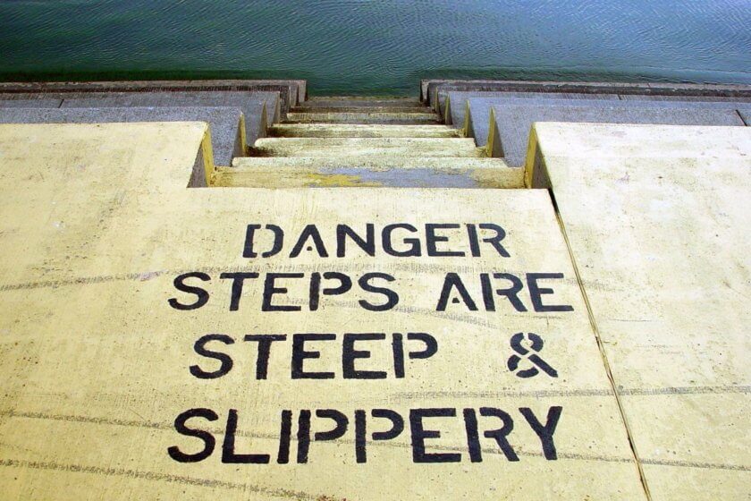Scheinselbstständigkeit / Betontreppe führt ins Wasser, auf dem Boden steht in schwarzer Schrift: Danger - Steps are steep and slippery
