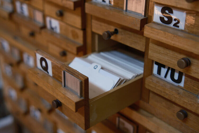 Klassischer Holz-Karteikasten mit alphabetisch sortierten Schubladen, eine davon geöffnet mit sichtbaren Karteikarten – analoges Kundendaten-Management vor dem digitalen CRM.