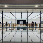 Arbeiten am Flughafen, Büro am Flughafen (Modernes, lichtdurchflutetes Bürogebäude mit großen Glasfassaden am Flughafen. Im Inneren sind Arbeitsstationen und einige Personen zu sehen, die an den Tischen arbeiten.)