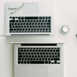 nebenberuflich selbstständig (Zwei Laptops auf einem weißen Schreibtisch, umgeben von einer Kerze, einer kleinen Pflanze, einem Notizbuch und einer Schale mit Keksen.)