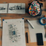 Immobilienunternehmer / Immobilien (Holztisch mit Skizzenbüchern, Architekturzeichnungen, Markern, Klebeband, einem Smartphone und einem Laptop, das kreative Arbeiten darstellt.)