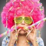 Weiberfastnacht / Frau mit pinker Lockenperücke, grüner Partybrille und bunten Luftschlangen, die in zwei Partytröten bläst.