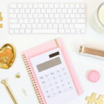 Kostenreduktion / Schreibtisch mit weißer Tastatur, Taschenrechner, Notizbuch und goldenen Büroaccessoires – Symbole für Finanzplanung und Kostenkontrolle.