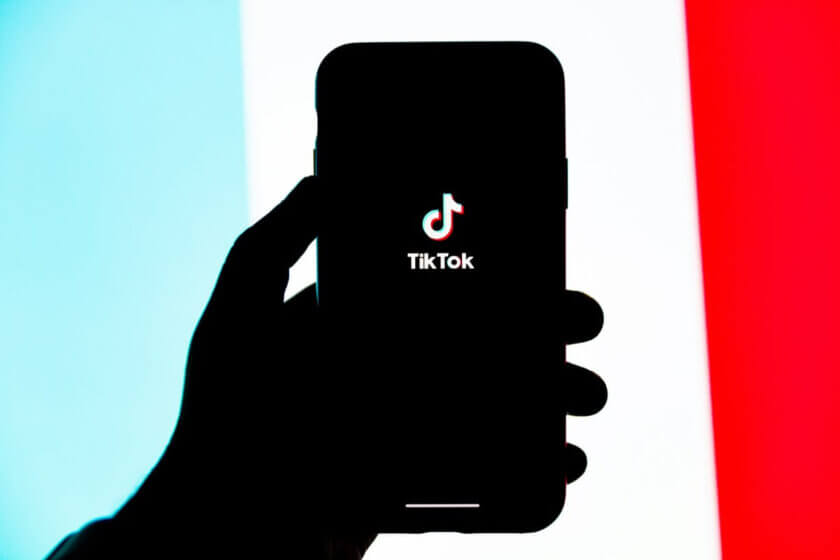 TikTok Follower kaufen (Hand hält ein Smartphone mit geöffnetem TikTok-Startbildschirm vor einem Hintergrund in Hellblau, Weiß und Rot.)