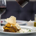 Restaurant (Lasagne mit Sahnesoße und Käsechip auf einem weißen Teller, frisch gepfeffert – im Hintergrund ein Glas Rotwein, Ölflasche und stilvolles Tischgedeck.)