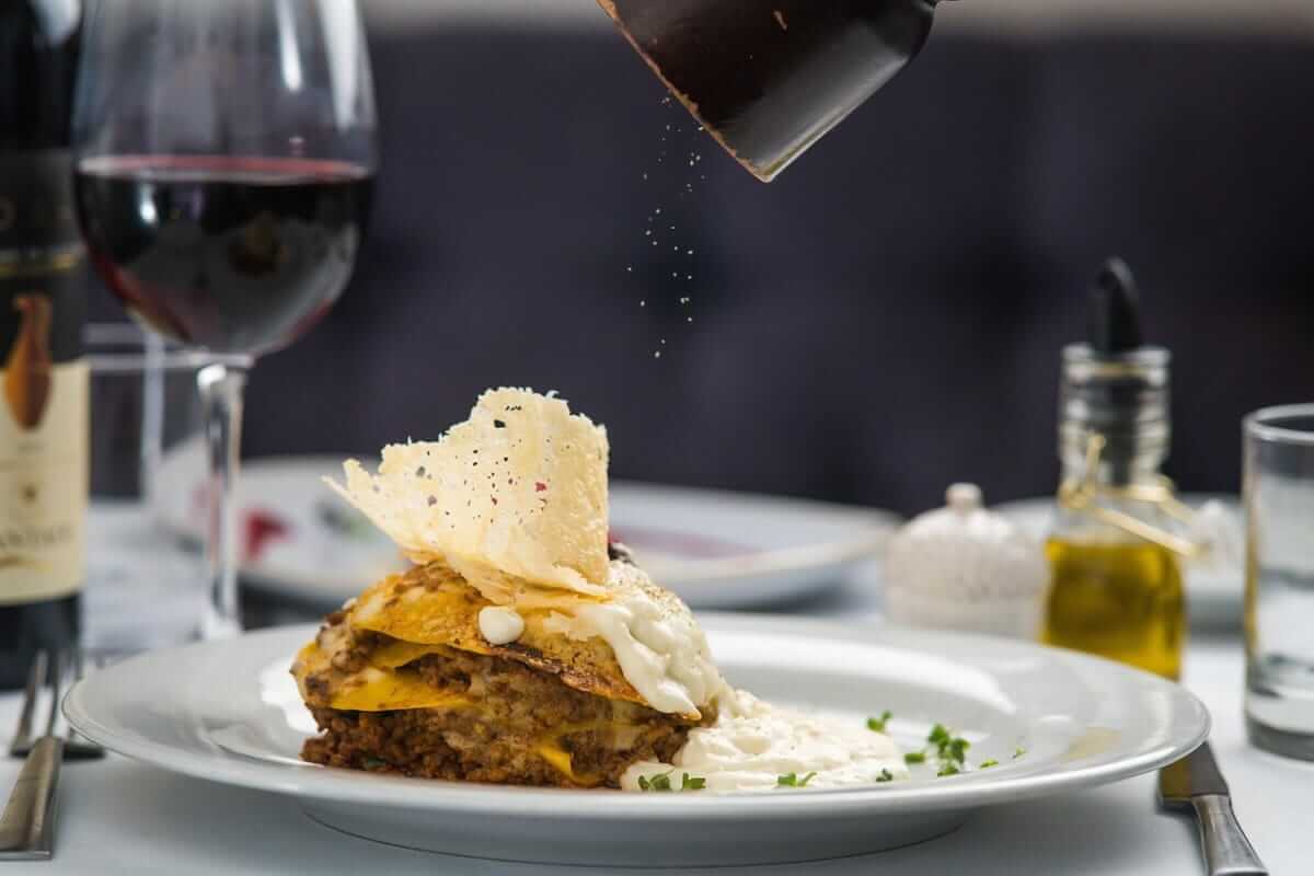 Restaurant (Lasagne mit Sahnesoße und Käsechip auf einem weißen Teller, frisch gepfeffert – im Hintergrund ein Glas Rotwein, Ölflasche und stilvolles Tischgedeck.)