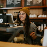 Minijob, Aushilfe / Junge Frau arbeitet als Aushilfe in einem Café und bedient die Kaffeemaschine mit einem Lächeln.