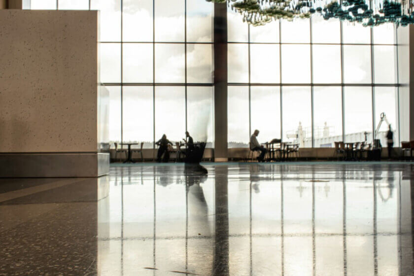 Büro am Airport / Ruhige Flughafenhalle mit großer Glasfront, vereinzelte Personen arbeiten oder warten an Tischen, sachliche und funktionale Arbeitsatmosphäre.