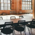 Leerer Konferenzraum mit schwarzen Stühlen und Holztisch, Licht fällt durch große Industriefenster – Symbol für überflüssige Meetings und verschwendete Zeit.