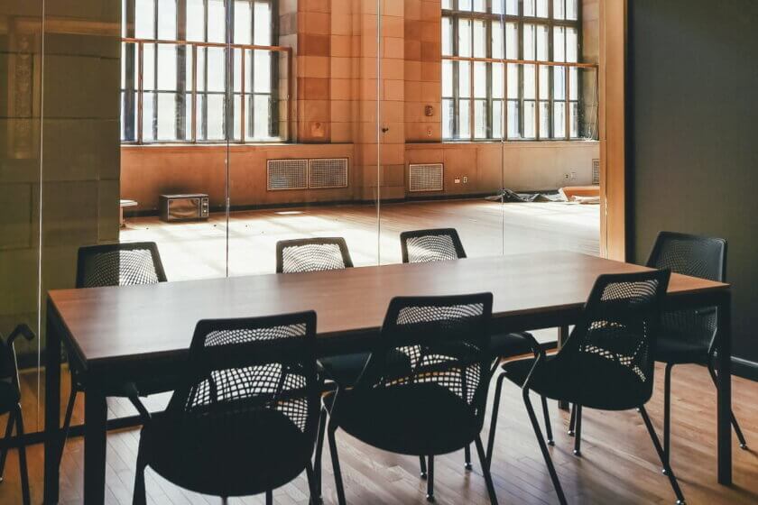 Leerer Konferenzraum mit schwarzen Stühlen und Holztisch, Licht fällt durch große Industriefenster – Symbol für überflüssige Meetings und verschwendete Zeit.