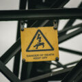 Kennzeichnungssysteme / Verwittertes gelbes Warnschild mit Blitzsymbol und Aufschrift „Danger of Death – Keep Off" an einer Stahlkonstruktion