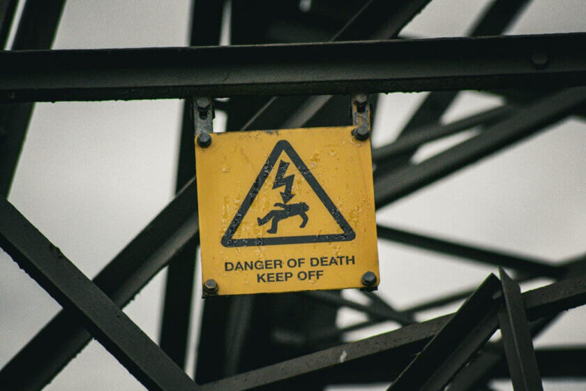 Kennzeichnungssysteme / Verwittertes gelbes Warnschild mit Blitzsymbol und Aufschrift „Danger of Death – Keep Off" an einer Stahlkonstruktion