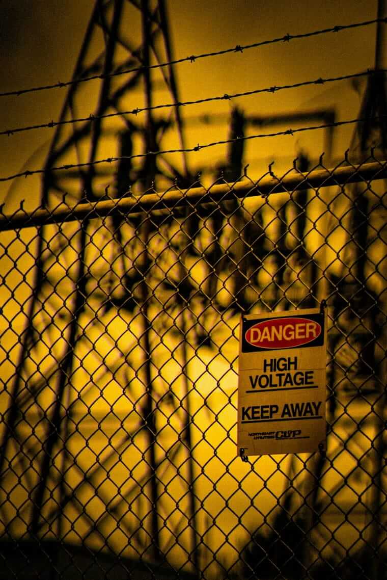 Kennzeichnungspflicht / Warnschild „Danger High Voltage – Keep Away" an einem Maschendrahtzaun vor einer Industrieanlage im Gegenlicht