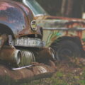 Zwei rostige Oldtimer auf einem Schrottplatz, am vorderen Fahrzeug ein Schild mit der Aufschrift „Rust in Peace" – Symbol für die Folgen falscher Investitionsentscheidungen.