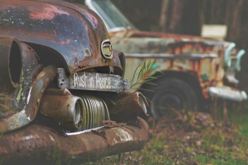Zwei rostige Oldtimer auf einem Schrottplatz, am vorderen Fahrzeug ein Schild mit der Aufschrift „Rust in Peace" – Symbol für die Folgen falscher Investitionsentscheidungen.