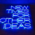 Preismodelle / Blaues Neonschild mit der Aufschrift „New Times for Other Ideas" an einer Backsteinwand – sinnbildlich für den Wandel in der Preisgestaltung und neue Monetarisierungsansätze.