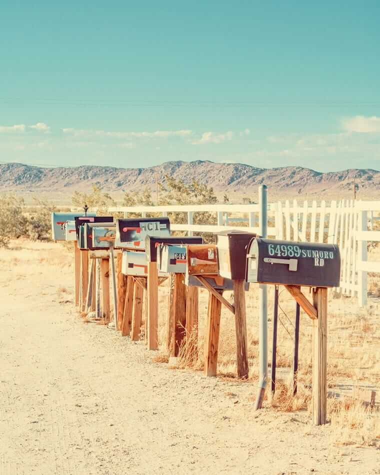 Newsletter / Eine Reihe alter Briefkästen steht auf Holzpfosten entlang einer staubigen Straße in der Wüste.