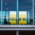 Silhouette einer Person mit Rollkoffer hinter einer Glasfront am Flughafen, im Hintergrund ein gelbes Schild mit der Aufschrift „Follow Me"