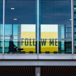 Silhouette einer Person mit Rollkoffer hinter einer Glasfront am Flughafen, im Hintergrund ein gelbes Schild mit der Aufschrift „Follow Me"
