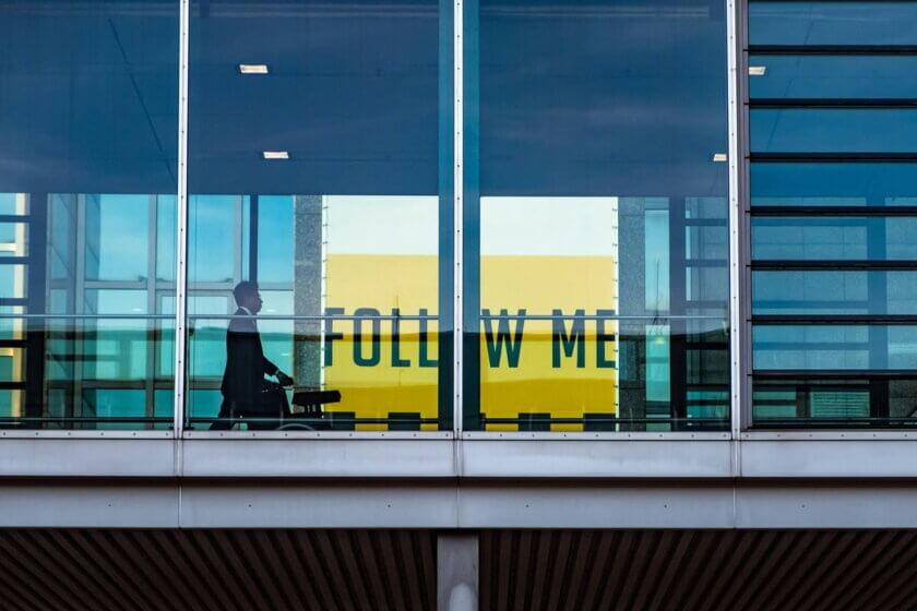 Silhouette einer Person mit Rollkoffer hinter einer Glasfront am Flughafen, im Hintergrund ein gelbes Schild mit der Aufschrift „Follow Me"