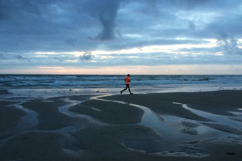 Einzelner Läufer am frühen Morgen am Strand bei bewölktem Himmel – Sinnbild für Disziplin und Ausdauer auch ohne Motivation.